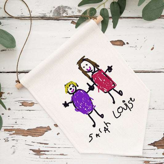 Children's drawing of two stick figures on a white sheet of paper with 'Sarah Louise' written below, placed on a wooden surface with green leaves. | Little Heirloom