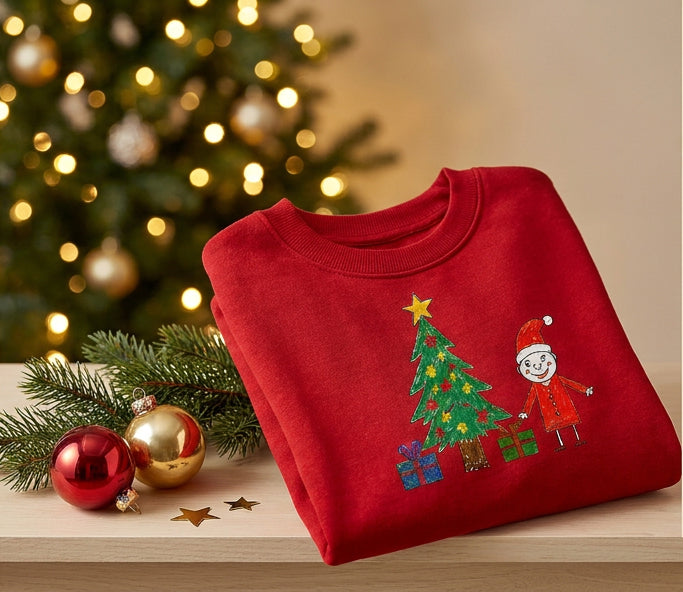 A folded red crewneck sweatshirt with a Christmas tree and Santa Claus graphic, styled on a wooden surface with a blurred Christmas tree, ornaments, and pine branches in the background.