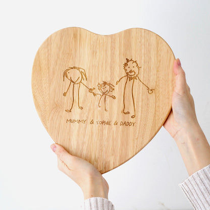 Heart-shaped wooden cutting board with engraved family illustration drawn by a child and names, held by a person.| Little Heirloom