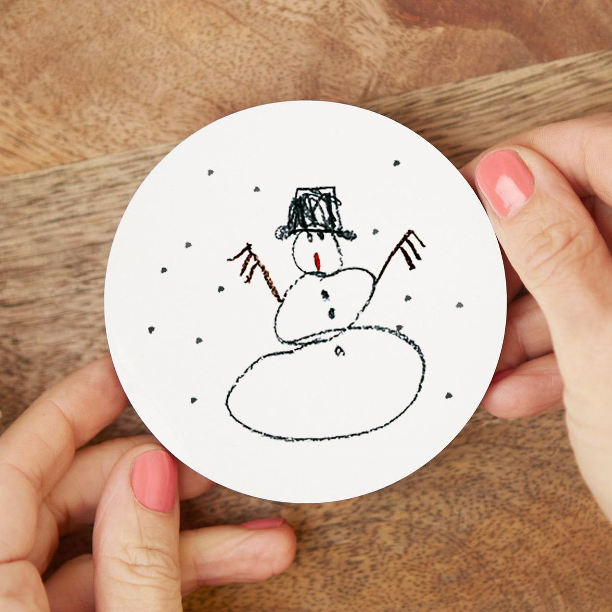 Hand holding a round white coaster with a snowman drawing on a wooden surface | Little Heirloom