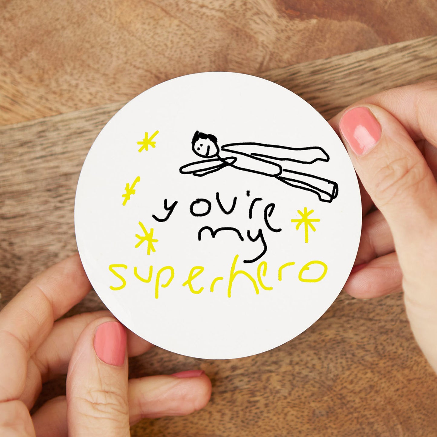 Circular sticker with 'You're my superhero' text and illustration held by hands on a wooden surface| Little Heirloom