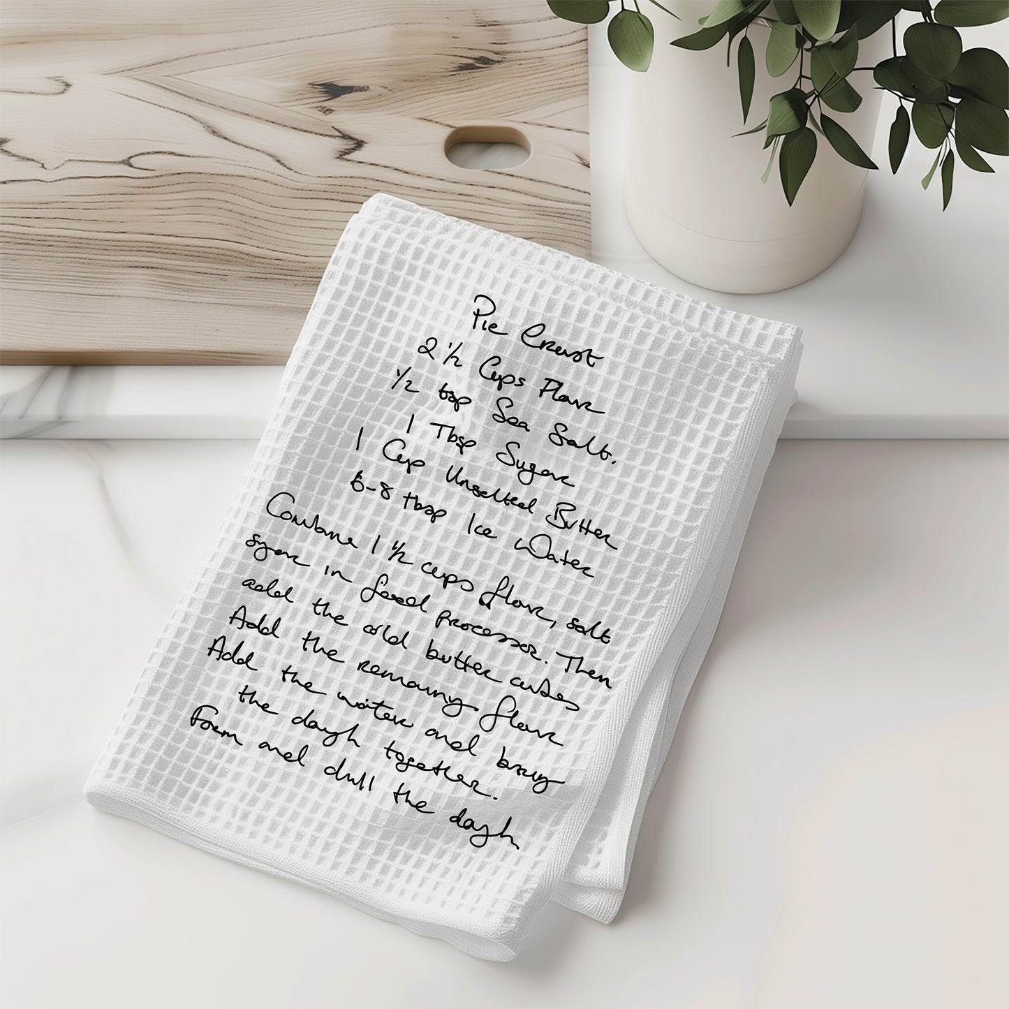 White kitchen towel with handwritten recipe on a wooden surface with a vase of greenery.| Little Heirloom