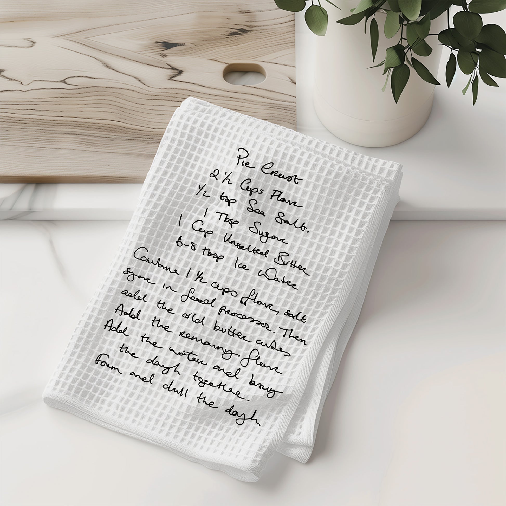 White kitchen towel with handwritten recipe on a wooden surface with a vase of greenery.| Little Heirloom