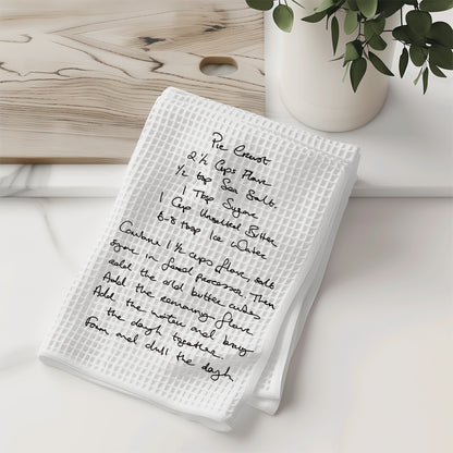 White kitchen towel with handwritten recipe on a wooden surface with a vase of greenery.| Little Heirloom