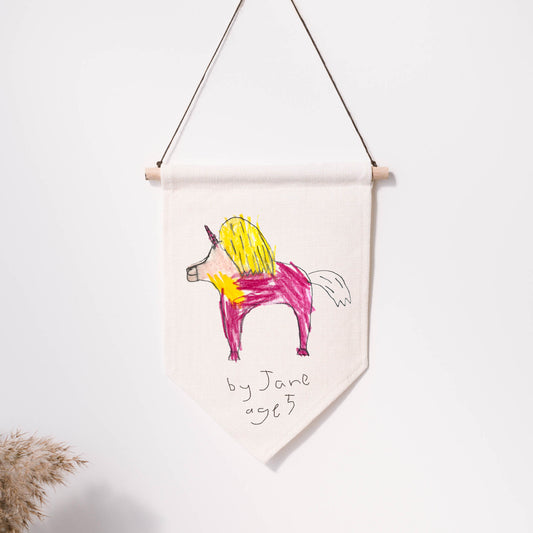 Hand-drawn unicorn on a fabric banner with 'by Jane 983' written below, hanging against a white wall. | Little Heirloom
